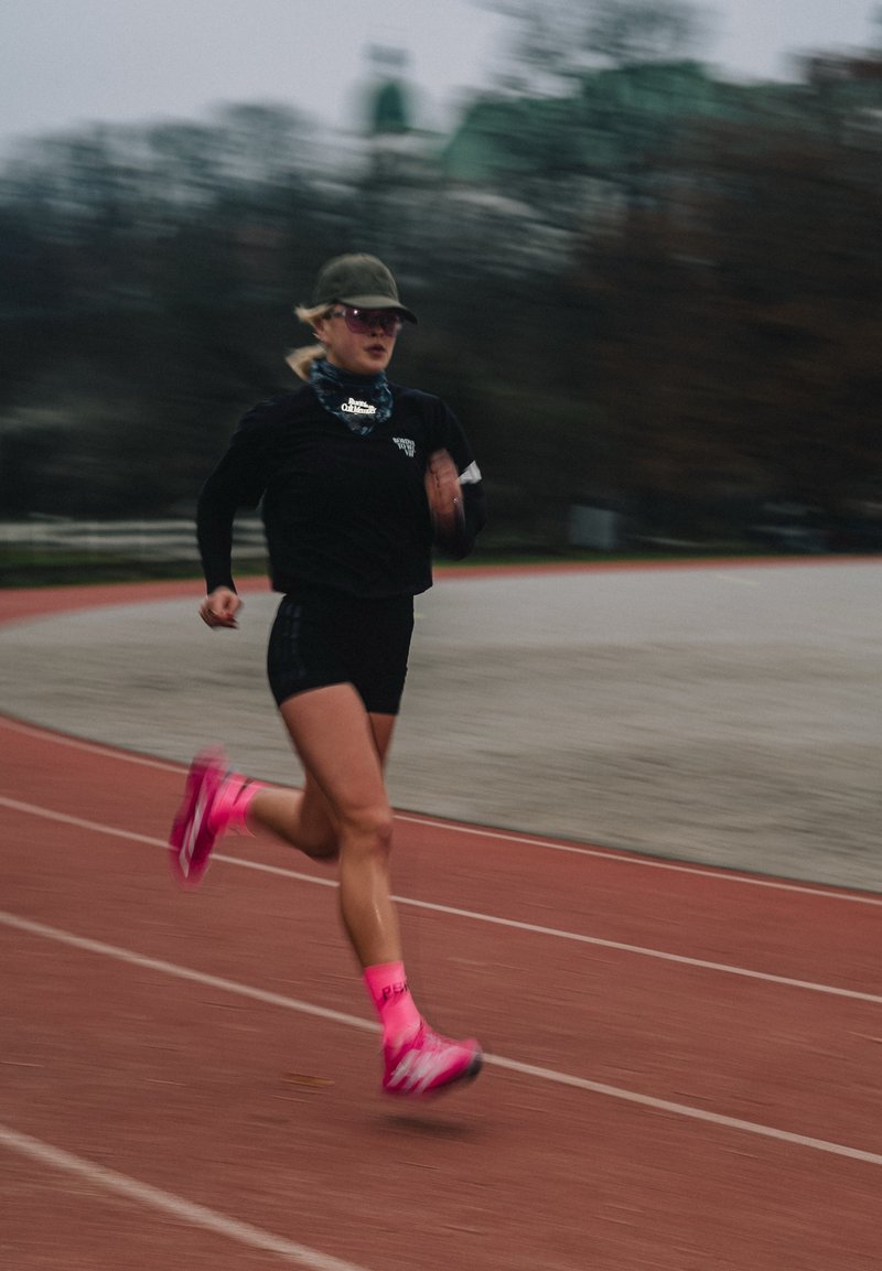 Hardloper in een zwart shirt met korte mouwen en zwarte korte broek, die roze sokken en felroze schoenen draagt, bewegend op een rode baan.