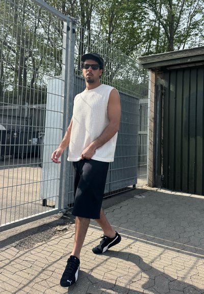 Top de punto sin mangas blanco, pantalones cortos negros a la rodilla y zapatillas negras. Se encuentra cerca de una valla y un edificio verde; la luz del sol crea sombras.