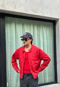 Κόκκινο νάιλον τζάκετ με στρογγυλή γιακά, φορεμένο πάνω από μια κόκκινη αμάνικη μπλούζα. Μαύρα τζιν και γυαλιά ηλίου, με φόντο ανοιχτοπράσινες κουρτίνες.