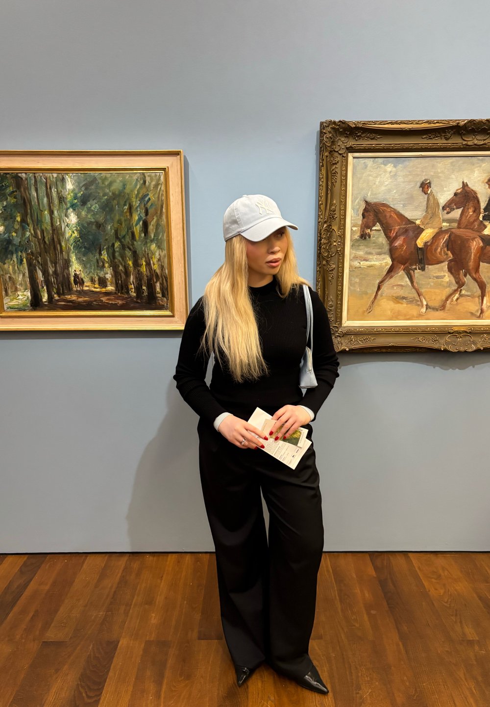 Jauna sieviete ar gariem blondiem matiem un baltu cepuri stāv mākslas galerijā starp divām rāmiem apvilktām gleznām, turot brošūras un ģērbusies melnā apģērbā.