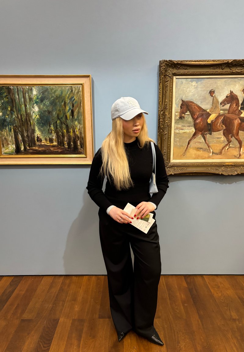 Mladá žena s dlouhými blond vlasy a bílou čepicí stojí v art galerii mezi dvěma rámovanými obrazy, drží brožury a má na sobě černé oblečení.