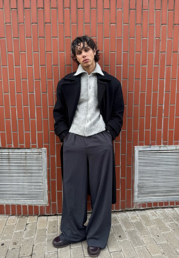 Νέος με σγουρά μαλλιά, που φοράει μαύρο παλτό, γκρι πουλόβερ, φαρδιά σκούρα παντελόνια και καφέ παπούτσια, στέκεται μπροστά σε έναν τοίχο από κόκκινα τούβλα.