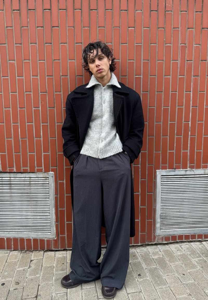 Νέος με σγουρά μαλλιά, που φοράει μαύρο παλτό, γκρι πουλόβερ, φαρδιά σκούρα παντελόνια και καφέ παπούτσια, στέκεται μπροστά σε έναν τοίχο από κόκκινα τούβλα.