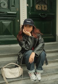 Black leather jacket, dark blue denim, beige handbag, white trainers with red accents, and a navy cap with the "Carhartt" logo. Seated on stone steps.