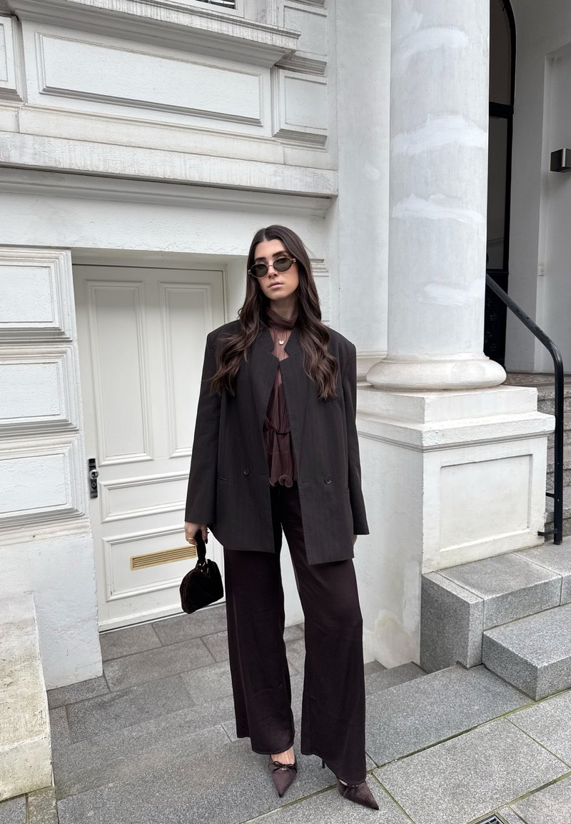 Femme portant un blazer noir surdimensionné, un pantalon à jambes larges, un haut transparent sombre, des lunettes de soleil, tenant un petit sac à main noir, debout près d'une colonne de bâtiment blanche.