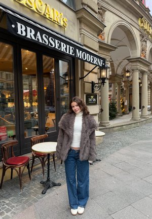 Abrigo de piel sintética marrón sobre un jersey de cuello alto blanco, combinado con jeans azules de pierna ancha y zapatillas blancas, frente a un restaurante con terraza al aire libre.