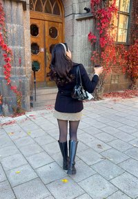 Zwarte jas over een geruite rok, doorschijnende zwarte panty, en zwarte laarzen tot de knie. Houdt een koffie vast dichtbij een stenen gebouw met klimop.