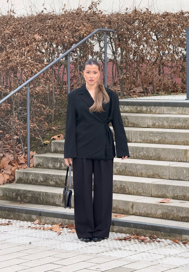 Blazer ajustado preto com mangas longas e detalhe de laço, emparelhado com calças largas pretas, e segurando uma pequena mala preta.