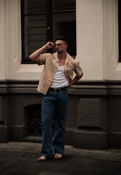 Camisa de rayas beige de manga corta, llevada sobre una camiseta blanca sin mangas, combinada con jeans acampanados azules y zapatos marrón y blancos, apoyada contra una pared.