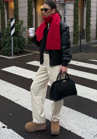 Chaqueta de cuero negra, bufanda roja de punto, pantalones de pana color crema, botas plataforma color camel, bolso negro. De pie en un paso de peatones sosteniendo una taza de café.