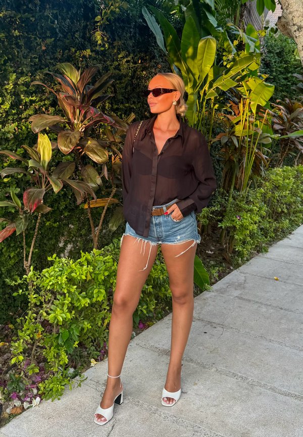 Una mujer con gafas de sol, una blusa negra, pantalones cortos vaqueros y zapatos blancos de tacón está de pie junto a exuberantes plantas tropicales en una acera.