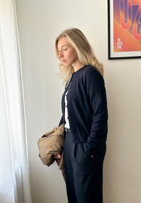 Cardigan marine en tricot doux avec des boutons, porté sur un haut blanc. Vêtement beige tenu d'un bras, pantalon noir, fond clair.