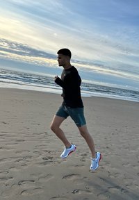 Un coureur en haut à manches longues noir et en shorts verts, portant des baskets blanches et rouges, fait du jogging sur une plage de sable avec des vagues de l'océan en arrière-plan.