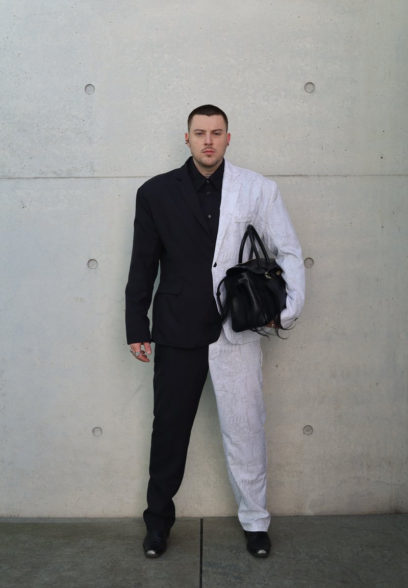Split suit: left side black, right side patterned white, textured fabric. Holds a black handbag. Concrete wall background.