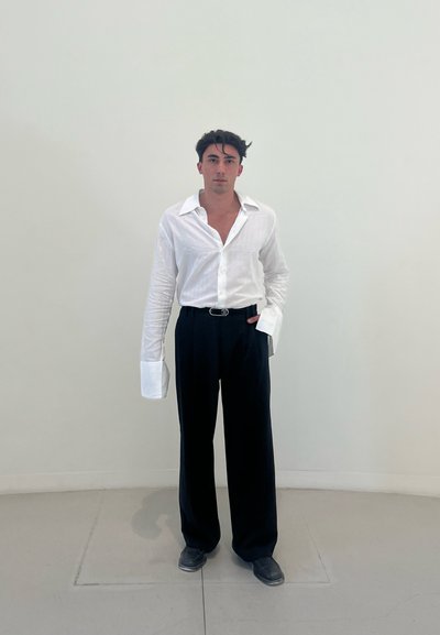 Camisa blanca de mangas largas con un patrón texturizado; puños de gran tamaño. Pantalones anchos negros con rayas verticales. Zapatos oscuros. Fondo liso.
