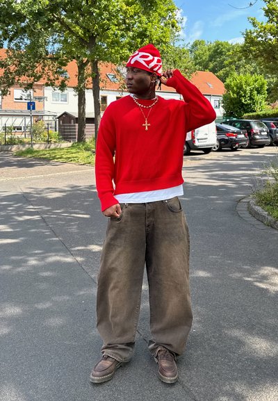 Suéter rojo con rayas sobre una camisa blanca, pantalones anchos marrones y botas marrones. Un gorro rojo y blanco con letras y un collar con una cruz.