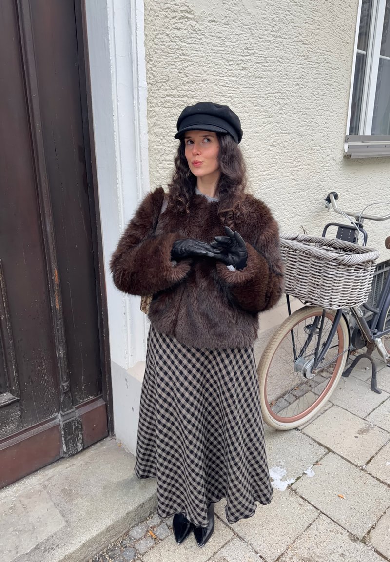 Veste en fausse fourrure marron, gants noirs, haut gris, jupe à carreaux noirs, chapeau noir. Vélo avec panier tressé en arrière-plan contre un mur texturé.