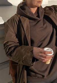 Casaco oversized castanho sobre um hoodie escuro, com uma mão segurando uma chávena de café estampada. Texturas suaves e estilo casual.
