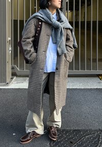 Personne portant un long manteau à carreaux, une chemise bleu clair, un pantalon beige, des bottes marron, un pull gris noué autour des épaules, debout les mains dans les poches.