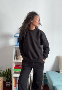 Vrouw met krullend haar, gekleed in een zwarte sweatshirt en broek, staat met haar handen in haar zakken en kijkt naar de zijkant in een kamer vol boeken en planten.