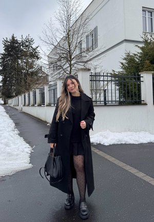 Joven mujer de cabello largo que lleva un abrigo negro, falda y medias de lunares, sosteniendo un bolso negro en una calle nevada junto a edificios blancos.