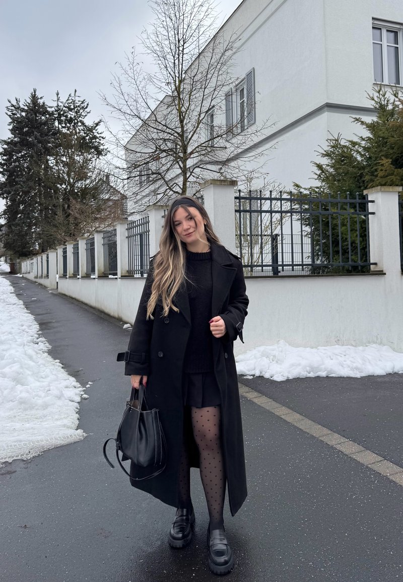 Jeune femme aux longs cheveux portant un manteau noir, une jupe et des collants à pois, tenant un sac à main noir dans une rue enneigée près de bâtiments blancs.