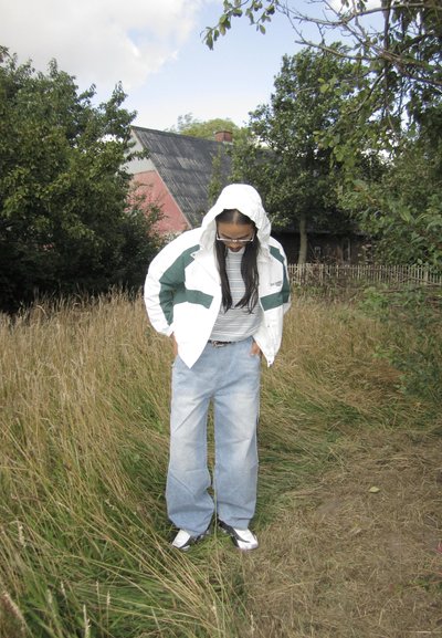 Chaqueta blanca y verde con un patrón y capucha, camiseta gris con rayas, pantalones vaqueros anchos de color azul claro, y zapatillas deportivas, de pie en hierba alta.