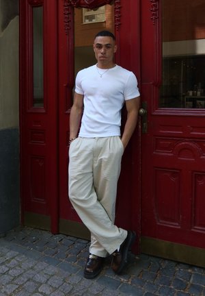 Camiseta blanca ajustada, pantalones sueltos beige y zapatos marrones con acentos dorados; está de pie contra una puerta de madera roja con detalles ornamentales.