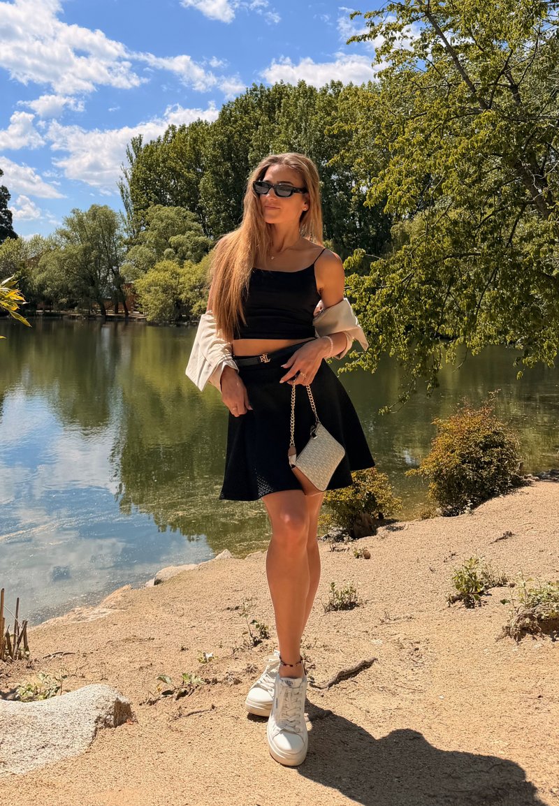Γυναίκα με μαύρο crop top, φούστα, άσπρα αθλητικά παπούτσια, γυαλιά ηλίου και κρατώντας μια μικρή τσάντα στέκεται δίπλα σε μια λίμνη με δέντρα κάτω από μπλε ουρανό.