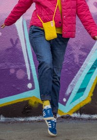 Ροζ επενδυμένο μπουφάν, κίτρινη πλεκτή τσάντα, μπλε τζιν με ελαστικούς ιμάντες και μπλε αθλητικά παπούτσια με λευκές ρίγες, μπροστά από έναν πολύχρωμο τοίχο γκράφιτι.