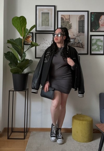 Chaqueta varsity negra con rayas blancas, vestido bodycon gris, bolso negro y zapatillas altas negras. Planta verde y arte enmarcado al fondo.