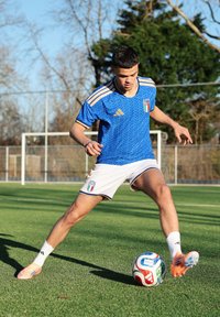 Νέος άνδρας ποδοσφαιριστής με μπλε φανέλα της Ιταλίας ελέγχει την μπάλα σε χλοοτάπητα, με τέρμα και δέντρα στο παρασκήνιο.