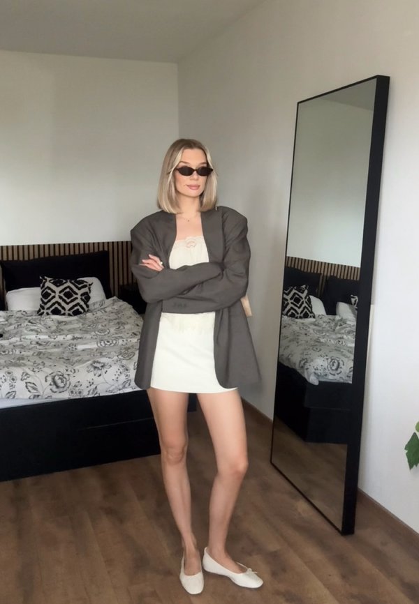 Femme portant des lunettes de soleil, une robe blanche et un blazer gris, debout les bras croisés dans une chambre avec un lit à motifs et un grand miroir.