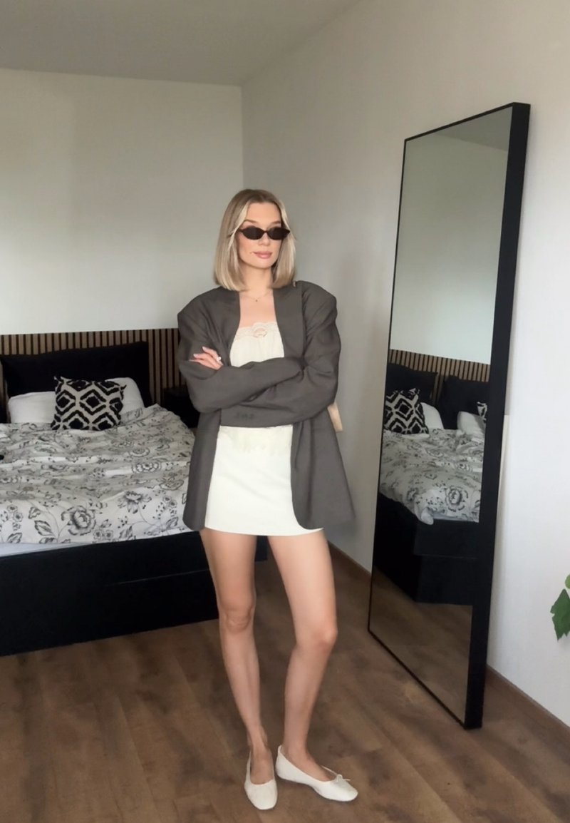 Femme portant des lunettes de soleil, une robe blanche et un blazer gris, debout les bras croisés dans une chambre avec un lit à motifs et un grand miroir.