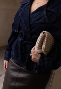 Mørk denimjakke med bælte i taljen, parret med en brun læderskirt. Holder en vævet beige cluthar. Enkel, struktureret design.