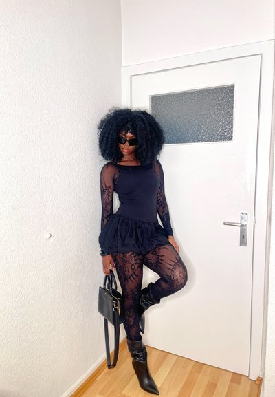 Bodysuit negro de manga larga con dobladillo con volantes, combinado con leggings negros de patrón de encaje y botas de tacón altas hasta la rodilla. Los accesorios incluyen un bolso negro.