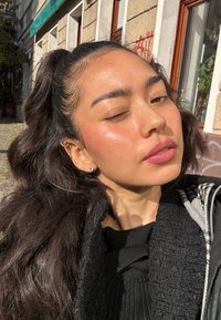 Zbliżenie na osobę z długimi, falującymi czarnymi włosami, noszącą czarną, wzorzystą bluzkę i srebrne kolczyki, z naturalną, promienną skórą.