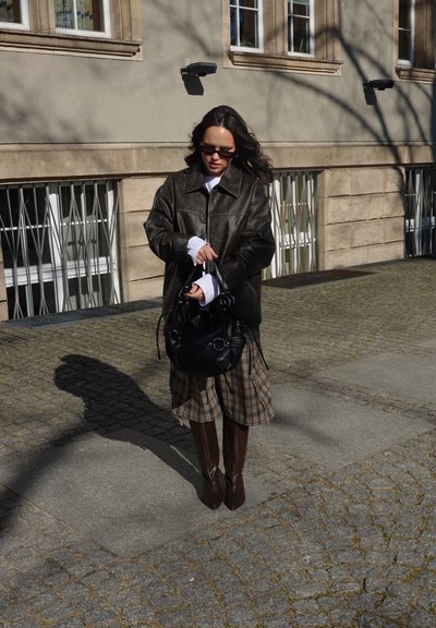 Mujer con chaqueta de cuero oscura, falda de cuadros y botas hasta la rodilla, sosteniendo un bolso negro, parada en una calle empedrada iluminada por el sol junto a un edificio.