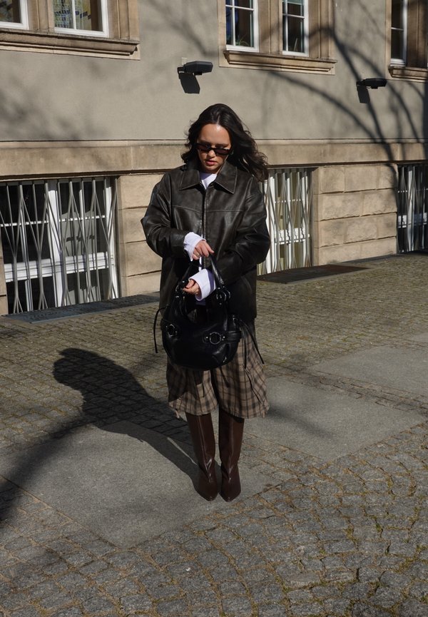 Femeie purtând o jachetă de piele neagră, fustă în carouri și cizme până la genunchi, ținând o geantă neagră, stând pe o stradă pietruită luminată de soare lângă o clădire.