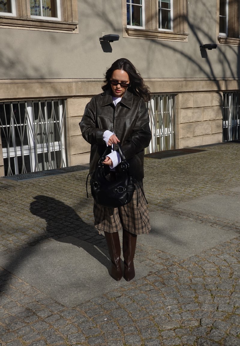 Vrouw draagt een donker leren jack, geruite rok en kniehoge laarzen, houdt een zwarte handtas vast en staat op een zonovergoten straat met keien bij een gebouw.