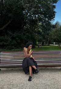 Drewniana ławka z metalowymi akcentami. Osoba w jasnoniebieskiej bezrękawnikowej bluzce i czarnej plisowanej spódnicy siedzi na zewnątrz w parku.