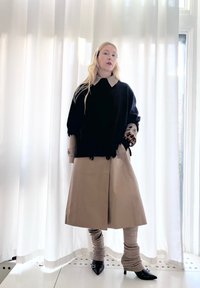 Woman with long blonde hair wearing a black jumper over a beige trench coat, beige leg warmers, and black pointed heels standing indoors by white curtains.