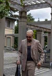 Ανοιχτό καφέ σουέτ κοστούμι με διπλό μπουφάν και ταιριαστά παντελόνια, φορεμένο πάνω από μαύρο τιραντάκι; κρατώντας μια μαύρη τσάντα.