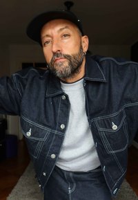 Donkerblauwe denimjas met contrasterende witte stiksels, uitgerust met twee borstzakken en zilveren hardware; gedragen over een grijze shirt.