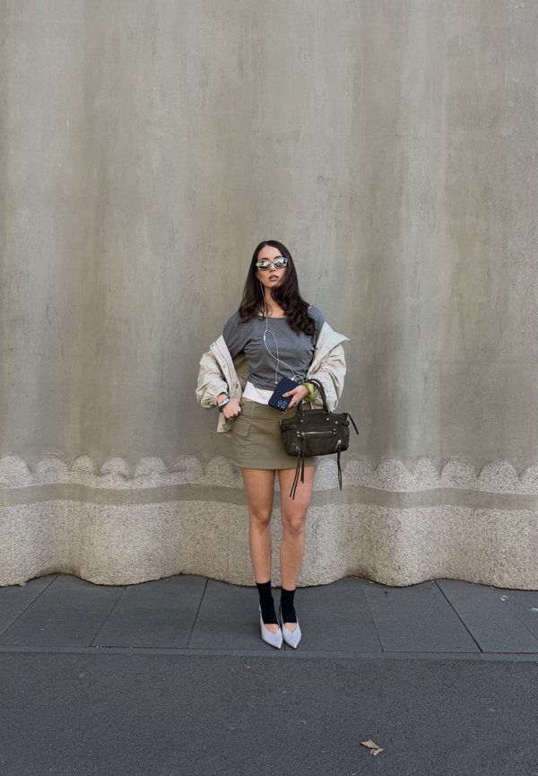 Une femme portant des lunettes de soleil pose devant un mur en béton. Elle porte un haut gris, une jupe kaki et une veste légère, et tient un sac à main sombre ainsi qu’un téléphone.