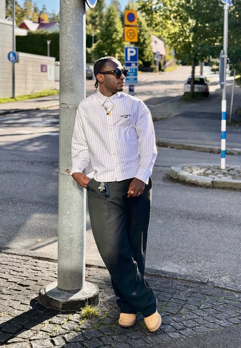 White striped button-up shirt, black loose-fitting pants, and light brown clogs. Accessories include layered gold necklaces and sunglasses.