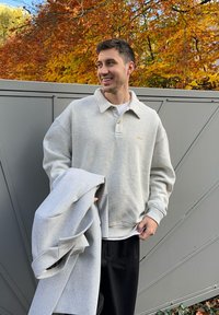 Sweatshirt cinzento claro com gola dobrada e três botões, apresenta um pequeno logótipo dourado. Segura um blazer cinzento claro; ao fundo, folhas de outono.