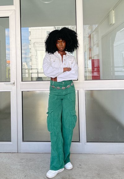 Chaqueta de mezclilla blanca, pantalones cargo de pana verde, tejido texturizado, cinturón con herrajes plateados, de pie frente a un fondo de puerta de vidrio.