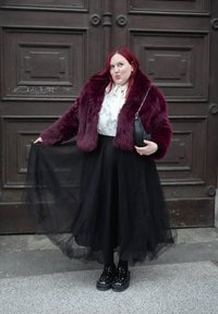 Γυναίκα με κόκκινα μαλλιά που φοράει μπέρντι πανωφόρι από γούνα, λευκό πουκάμισο και μαύρη φούστα από τούλι, κρατώντας τη φούστα της και μια μαύρη τσάντα χειρός.