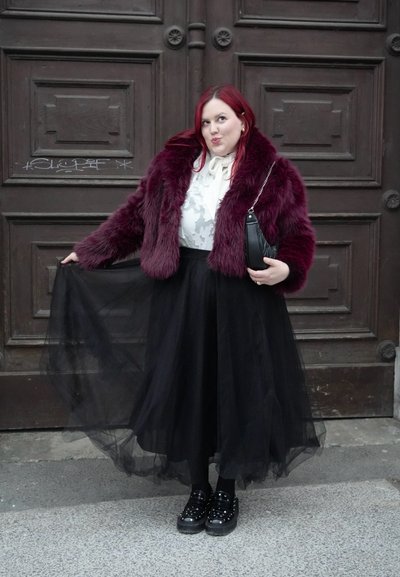 Mujer con cabello rojo, vestida con una chaqueta de piel burdeos, blusa blanca y falda de tul negra, sosteniendo su falda y un bolso negro.